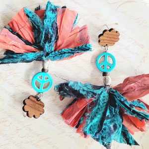 May include: A pair of dangle earrings with turquoise peace sign charms and wooden cloud charms. The earrings are made with red and turquoise fabric tassels.