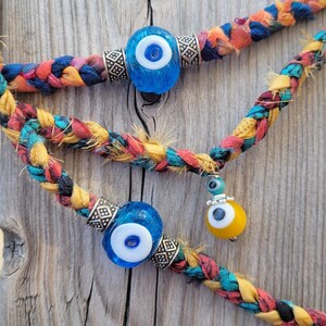 May include: A braided cord necklace with three blue evil eye beads and one yellow evil eye bead. The cord is made of colorful fabric and is braided together.