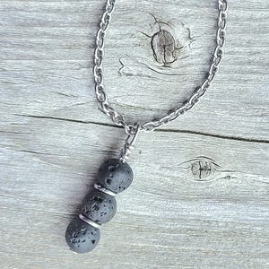 May include: A silver chain necklace with three dark gray lava stone beads. The beads are separated by silver rings and suspended from a silver chain. The necklace is displayed on a weathered wooden surface.