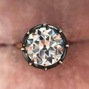 May include: Close-up of a large, clear diamond with a complex, multi-faceted cut. The diamond is set in a dark metal bezel with small, decorative prongs. The background is a soft, muted pink.