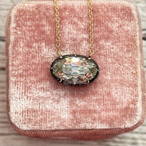 May include: A gold chain necklace with a large, oval-shaped, clear gemstone pendant set in a black metal bezel. The pendant is surrounded by small, black gemstones. The necklace is displayed in a pink velvet box. The text "@shanastamjewelry" is visible below the pendant.
