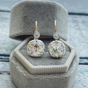 May include: A pair of gold and silver earrings with large, faceted, clear gemstones. Each earring has a smaller, round gemstone above the larger stone. The earrings are displayed in a gray velvet jewelry box.