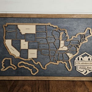 May include: A wooden map of the United States with each state outlined. The map is mounted on a dark gray background and framed in a brown wood frame. The text "Let the adventures begin" and "Travel Map" is printed on a wooden sign in the lower right corner of the map.