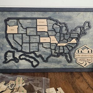 May include: A wooden US map with a distressed blue background. Each state is outlined in black and can be removed. The map is framed in black wood and has a sign at the bottom that says "Let the adventures begin Travel Map".