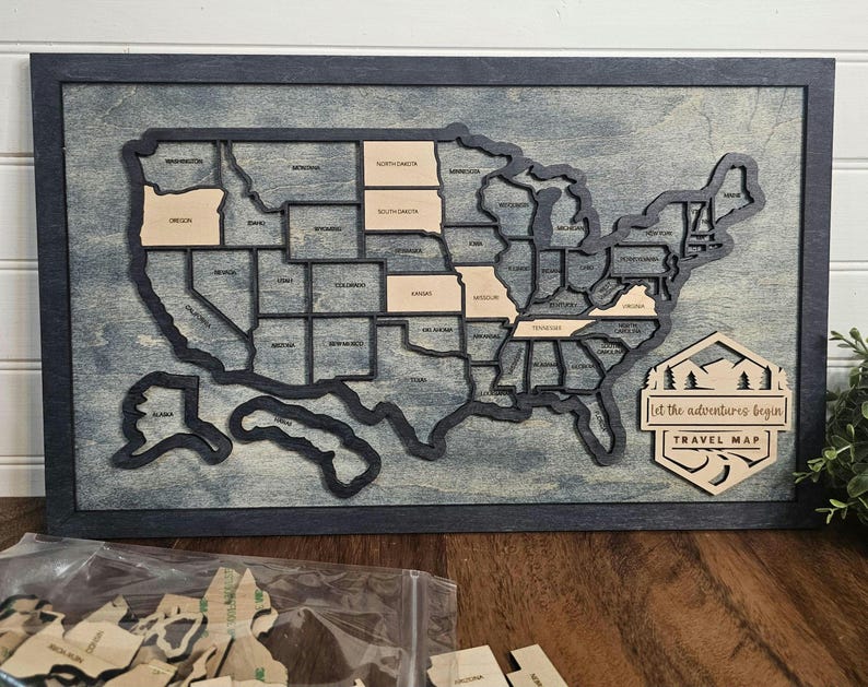 May include: A wooden US map with a distressed blue background. Each state is outlined in black and can be removed. The map is framed in black wood and has a sign at the bottom that says "Let the adventures begin Travel Map".