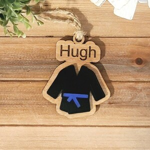 May include: Wooden ornament shaped like a karate uniform with a blue belt. The ornament has the name "Hugh" engraved on it.