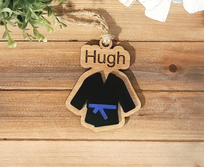 May include: Wooden ornament shaped like a karate uniform with a blue belt. The ornament has the name "Hugh" engraved on it.