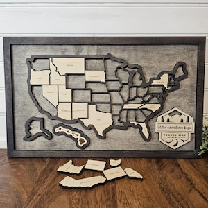 May include: A wooden map of the United States with each state as a separate piece. The map is framed in dark wood and has a gray background. The text "Let the adventures begin TRAVEL MAP" is on the bottom right corner of the map. The states of California, Oregon, and Washington are shown as separate pieces below the map.