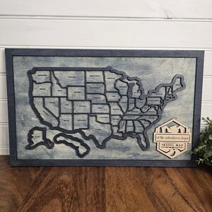 May include: A wooden map of the United States with each state outlined in black. The map is mounted on a dark blue frame and has a light blue background. The text "Let the adventures begin" and "Travel Map" is on a separate wooden piece in the lower right corner.