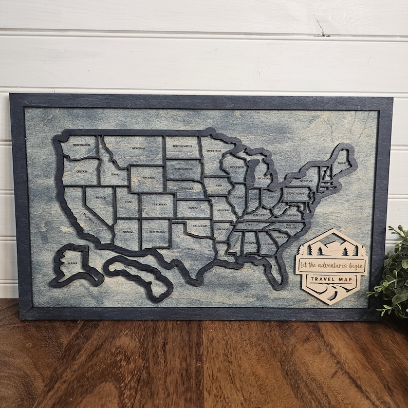 May include: A wooden map of the United States with each state outlined in black. The map is mounted on a dark blue frame and has a light blue background. The text "Let the adventures begin" and "Travel Map" is on a separate wooden piece in the lower right corner.