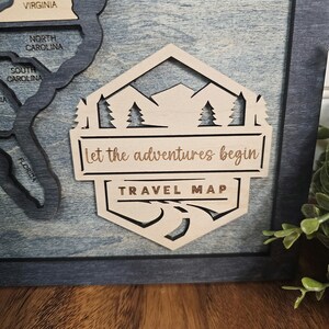 May include: A wooden travel map with a hexagonal design. The map features a mountain range, pine trees, and a winding road. The text "Let the adventures begin" and "TRAVEL MAP" is engraved on the map.
