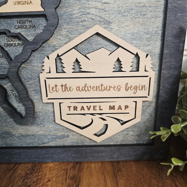 May include: A wooden travel map with a hexagonal design. The map features a mountain range, pine trees, and a winding road. The text "Let the adventures begin" and "TRAVEL MAP" is engraved on the map.