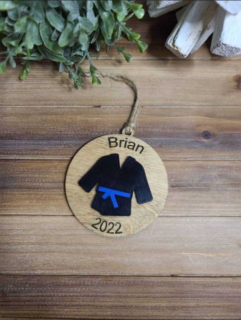 May include: A round wooden ornament with a black karate uniform and a blue belt. The ornament is personalized with the name "Brian" and the year "2022".