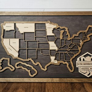 May include: Wooden travel map of the United States with state outlines and names. The map is framed in a dark wood with a lighter wood border. A decorative sign at the bottom reads "Let the adventures begin TRAVEL MAP."