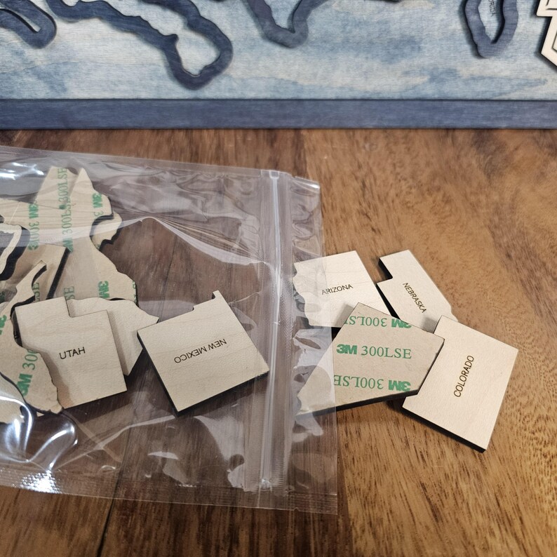 May include: A clear plastic bag containing several wooden state outlines, including Utah, New Mexico, Arizona, Nebraska, Colorado. The outlines are labelled with their respective state names and have adhesive backing.