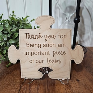 May include: A wooden puzzle piece with the text "Thank you for being such an important piece of our team." The puzzle piece is on a wooden stand.
