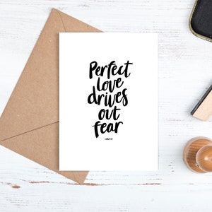 May include: A white card with black calligraphy that reads "Perfect love drives out fear". The card is sitting on a brown envelope on a white wooden surface.
