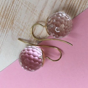 May include: A pair of faceted crystal ball earrings with gold-colored ear wires. The earrings are displayed on a pink and white wooden surface. The crystal balls have a geometric pattern.