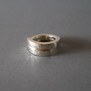 Könnte beinhalten: Ein silberner Ring mit gehammerter Textur. Der Ring hat ein einzigartiges Design mit zwei Bändern, die sich leicht überlappen. Der Ring wird vor einem grauen Hintergrund gezeigt.