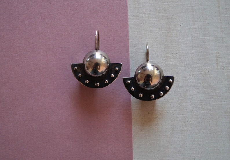 May include: A pair of silver earrings with a round, shiny, silver ball on top of a half-circle base with small silver dots around the edge.