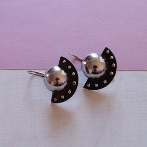 May include: A pair of silver earrings with a half-circle design and a round, reflective center. The earrings have a dotted pattern around the edge.