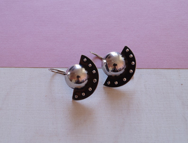 May include: A pair of silver earrings with a half-circle design and a round, reflective center. The earrings have a dotted pattern around the edge.