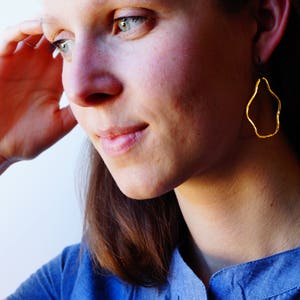 May include: A woman with long brown hair wearing a blue shirt and gold earrings with an irregular, organic shape.