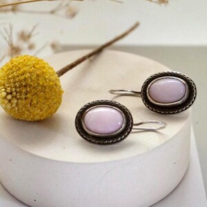 May include: A pair of silver-toned oval earrings with a light pink stone center. The earrings have a decorative border and a simple hook closure. A yellow flower and a white surface are in the background.