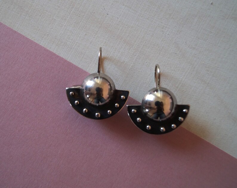 May include: A pair of silver earrings with a half-circle design and a round ball in the center. The earrings have a textured surface with small dots.
