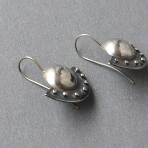 May include: A pair of silver earrings with a rounded, domed shape and a textured, slightly curved edge. The earrings have a small, silver hook.