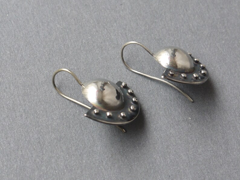 May include: A pair of silver earrings with a rounded, domed shape and a textured, slightly curved edge. The earrings have a small, silver hook.