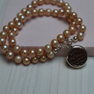 Puede incluir: Un collar de perlas de doble cadena con un dije de plata con un diseño grabado. Las perlas son de color melocotón claro.