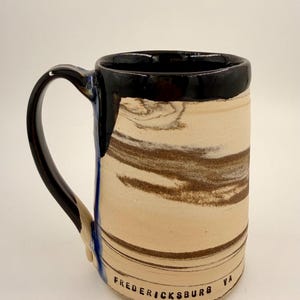 May include: A ceramic mug with a black rim and handle, featuring a beige and brown marbled design. The mug has the text "FREDERICKSBURG VA" printed on the side. The interior of the mug is black.