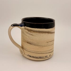 May include: A handmade ceramic mug with a cream-colored base and a dark brown and black rim. The mug has a curved handle and the text "FREDERICKSBURG VA" is printed on the side. The mug is approximately 4 inches tall.