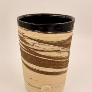 May include: A handmade ceramic tumbler with a unique design. The top rim is black, and the body features a swirling pattern of brown and cream tones. A small, embossed detail is visible near the base. This piece is ideal for beverages.