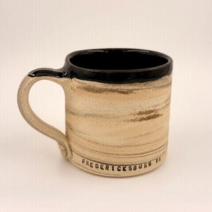 May include: A ceramic mug with a black rim and handle, and a beige and brown striped body. The text "FREDERICKSBURG VA" is printed on the side of the mug.
