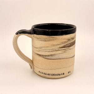 May include: A handmade ceramic mug with a black rim and handle. The body of the mug has a beige and brown marbled design. The text "Fredericksburg VA" is printed on the bottom of the mug.