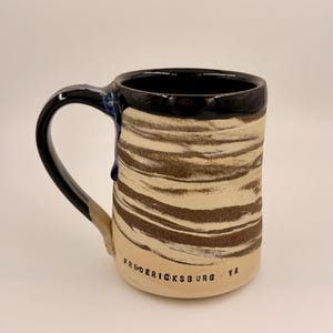 May include: A handmade ceramic mug with a black handle and rim. The mug features a beige and brown striped pattern on the body, with the text "Fredericksburg VA" printed on the base. A small blue glaze detail is visible.