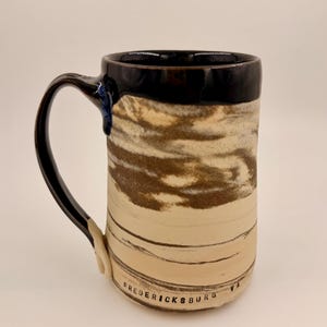 May include: A handmade ceramic mug with a black handle and rim. The body of the mug features a cream and brown marbled design. The text "FREDERICKSBURG VA" is printed on the bottom of the mug.