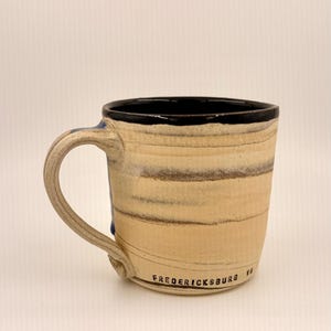 May include: A handmade ceramic mug with a beige and brown striped pattern. The rim is black, and the handle is beige. The text "FREDERICKSBURG VA" is printed on the bottom of the mug.