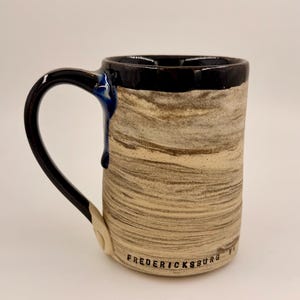 May include: A handmade ceramic mug with a black handle and rim, featuring a textured beige and brown body. The mug has a blue drip detail and the word "FREDERICKSBURG" is printed on the base.