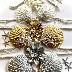 May include: A decorative arrangement featuring seashell bra tops in white, gold, and silver, adorned with faux pearls. The design includes floral accents, rope, and starfish earrings, creating a coastal theme.