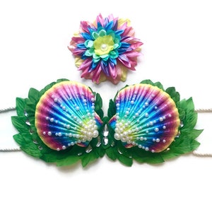 Könnte beinhalten: Ein regenbogenfarbener, schimmernder Muschel-BH mit Perlenimitationen und grünen Blättern. Ein passendes Blumenhaarband mit rosa, gelben, blauen und grünen Blütenblättern.