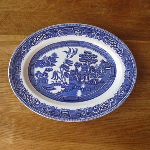 May include: Oval blue and white ceramic platter with a Willow pattern design. The platter features a detailed landscape scene with pagodas, trees, and a bridge. The rim has a decorative border. The platter is on a wooden surface.