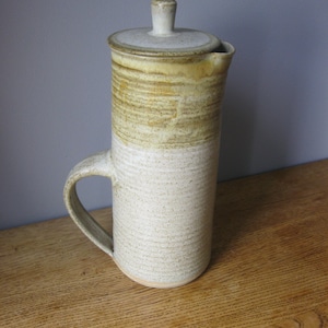 May include: A ceramic jug with a lid, featuring a brown and white glaze. The jug has a handle and a rounded body.