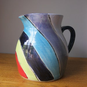 Può includere: Un'anfora in ceramica con un design a strisce colorate in tonalità di blu, verde, giallo, rosso e nero. L'anfora ha un manico e un'ampia apertura.