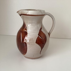 以下が含まれることがあります： クリーム色のベースと茶色の抽象的なデザインが施された、手作りのセラミックピッチャー。ピッチャーには、注ぎやすいように湾曲したハンドルと注ぎ口が付いています。デザインは有機的な形と線の組み合わせで、ユニークで素朴な美しさを生み出しています。