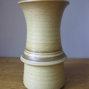 May include: A light brown ceramic vase with a wide mouth and a narrow base. The vase has a band of brown stripes around the middle.