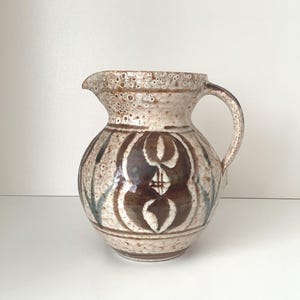 Puede incluir: Una jarra de cerámica con pico y asa. La jarra tiene un esmalte moteado blanquecino con diseños decorativos marrones y azules. El diseño presenta un motivo floral central y líneas verticales.