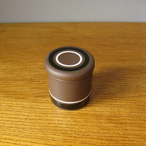 Könnte beinhalten: Eine braune Keramikdose mit einem schwarzen und weißen Kreisdesign auf dem Deckel.